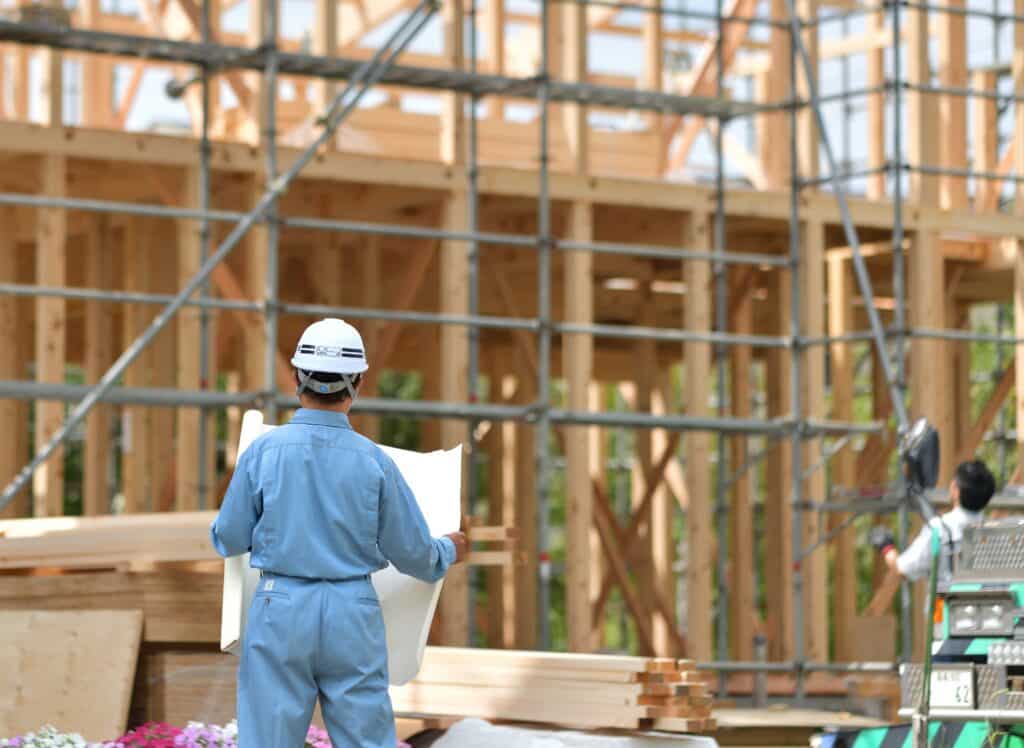Bauarbeiter mit Bauplan in der Hand vor einem Holzrohbau mit Gerüst