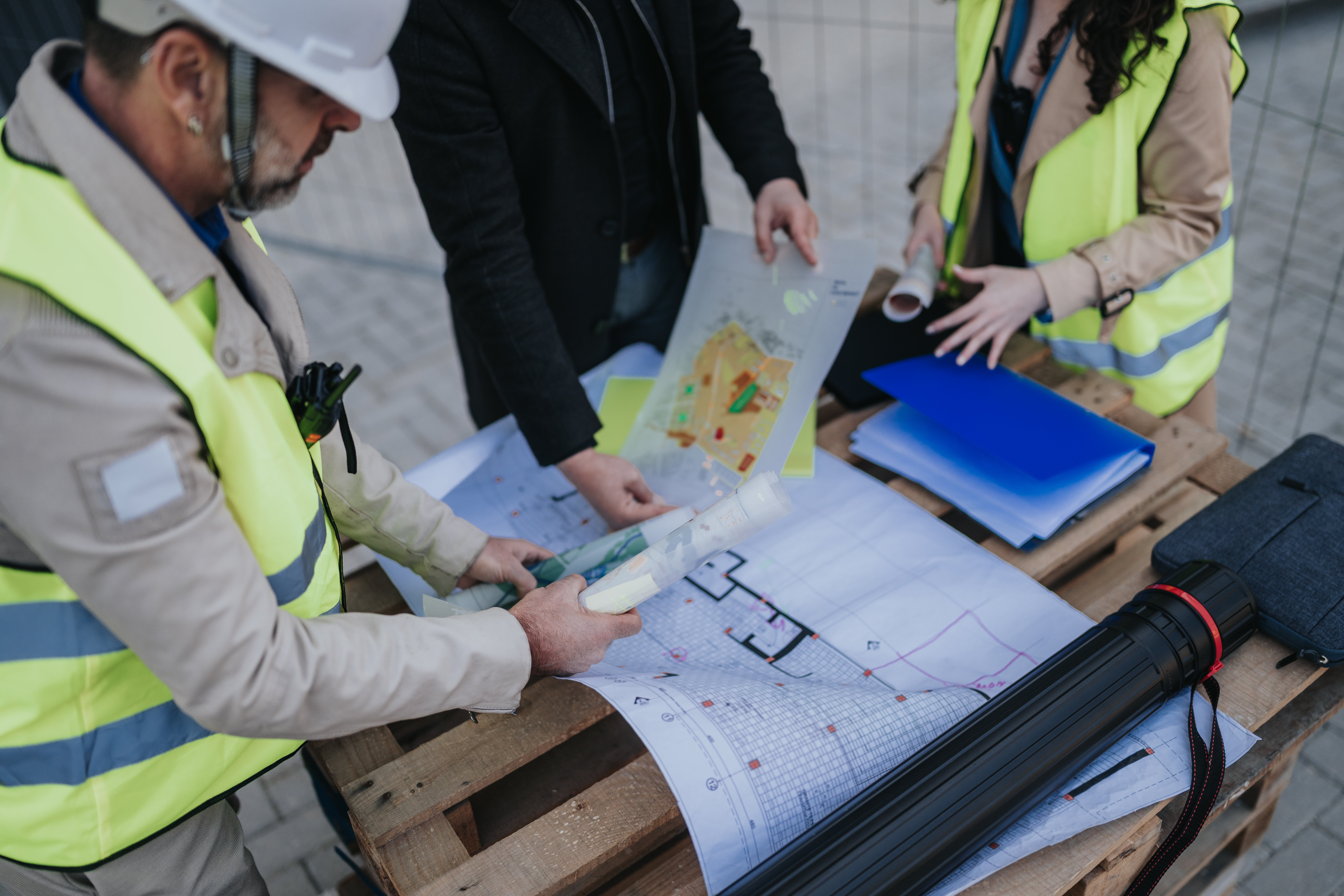 Drei Fachleute in Warnwesten und Schutzhelm besprechen auf einer Baustelle Baupläne und technische Zeichnungen