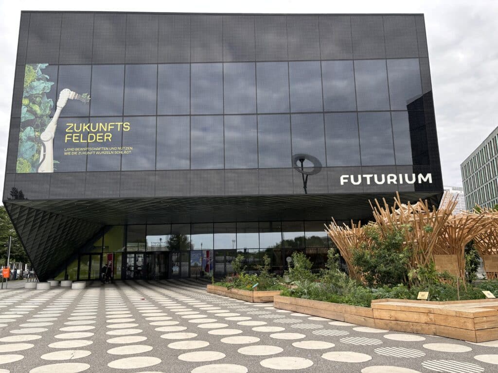 Außenansicht des Futuriums in Berlin mit dunkler Glasfassade, Schriftzug und bepflanzten Hochbeeten im Eingangsbereich